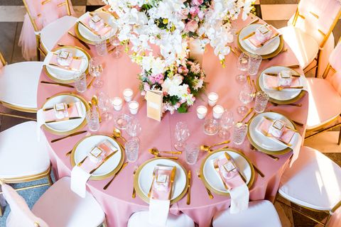 A round table with a pink tablecloth and gold-rimmed white plates, surrounded by pink chairs and centered with flowers. A round table with a pink tablecloth and gold-rimmed white plates, surrounded by pink chairs and centered with flowers.