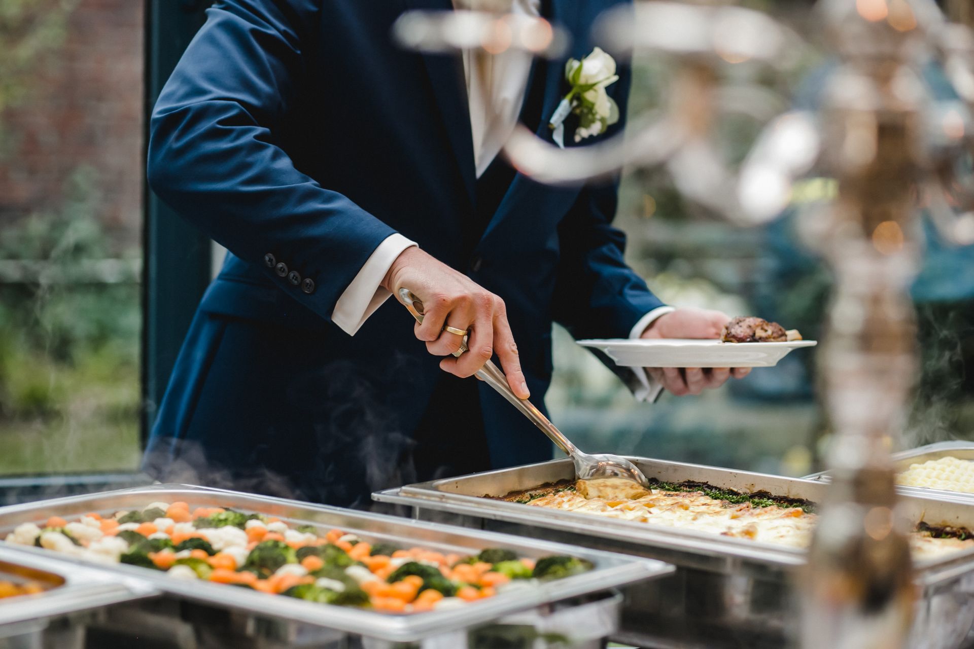A person in a blue suit uses serving tongs to place food onto a plate from a buffet of warm dishes.