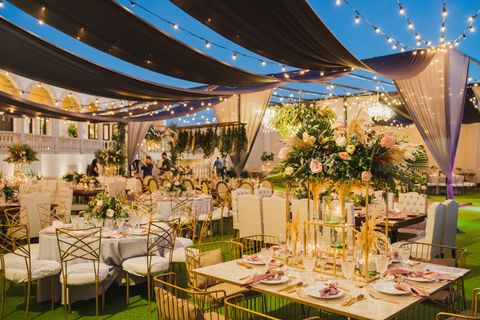 An outdoor evening banquet area with tables, gold chairs, and floral centerpieces under hanging fabric and string lights. An outdoor evening banquet area with tables, gold chairs, and floral centerpieces under hanging fabric and string lights.