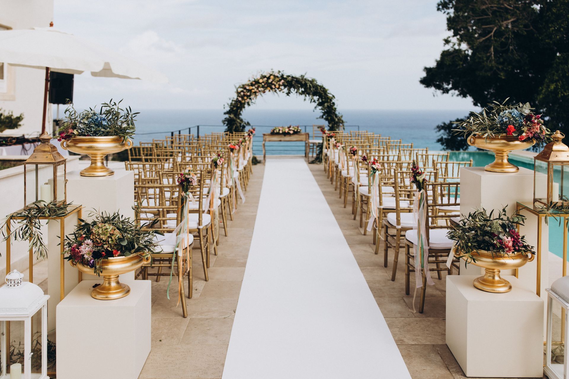 An outdoor wedding aisle lined with chairs, gold pedestals with floral arrangements, and a circular arch facing the ocean.