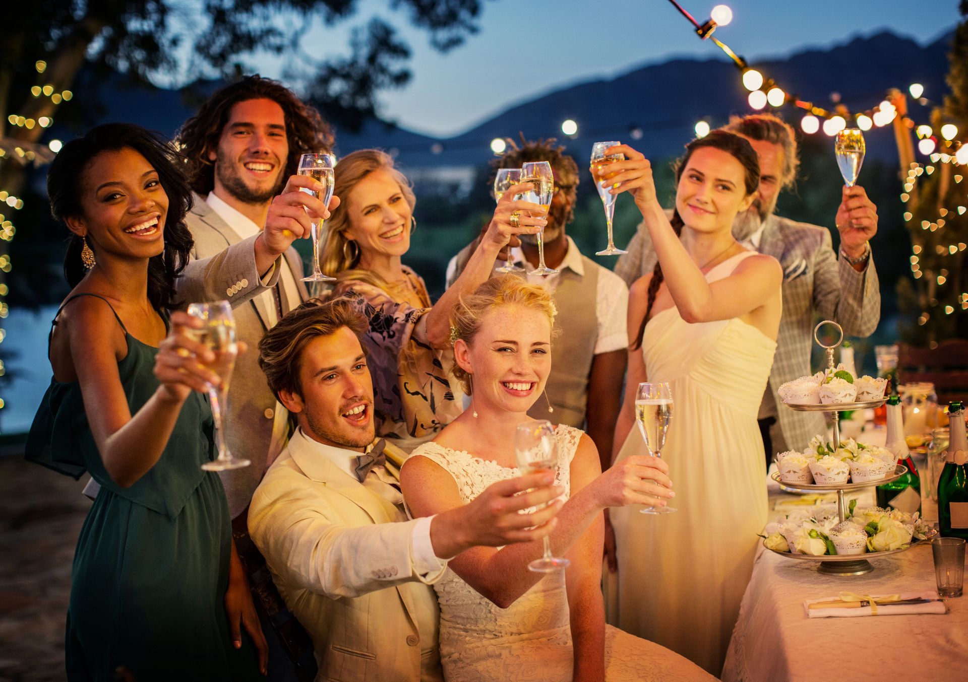 A group of friends in formal attire toast with champagne glasses at an outdoor evening event with string lights.