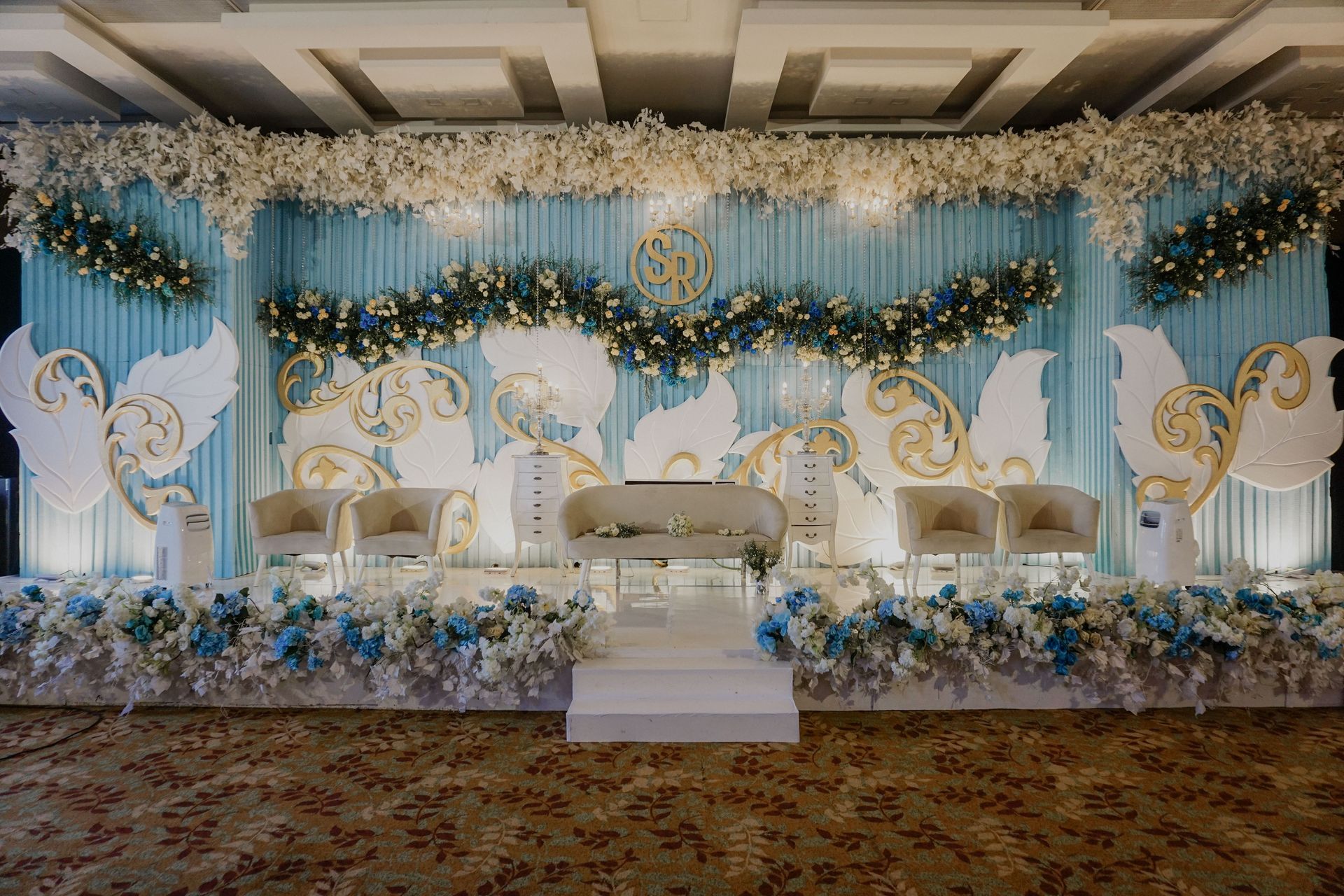 A wedding stage decorated with blue curtains, floral arrangements, golden accents, and white chairs on a raised platform.