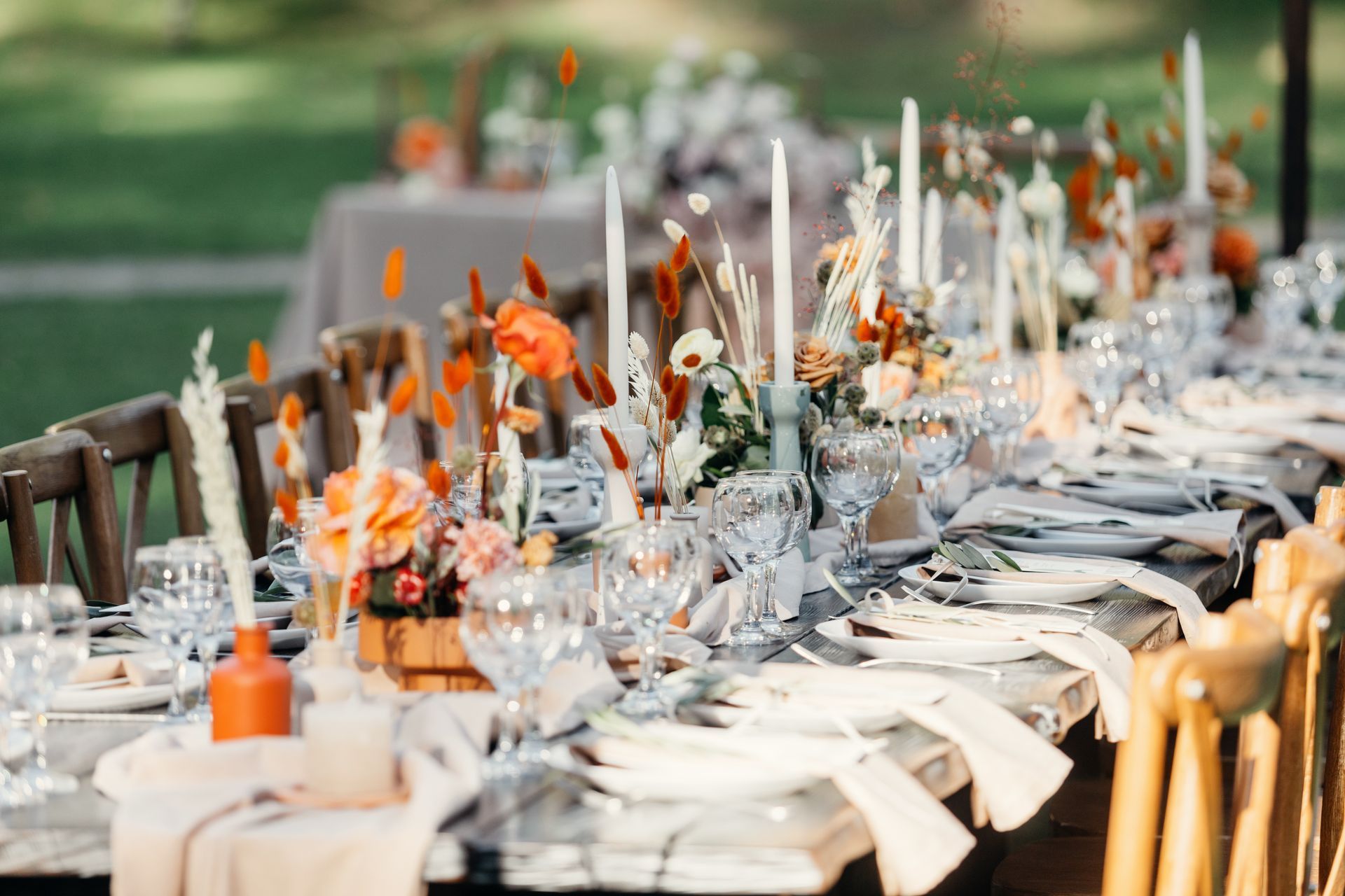 A long table set for an outdoor wedding with orange florals, white tapered candles, and light table linens.