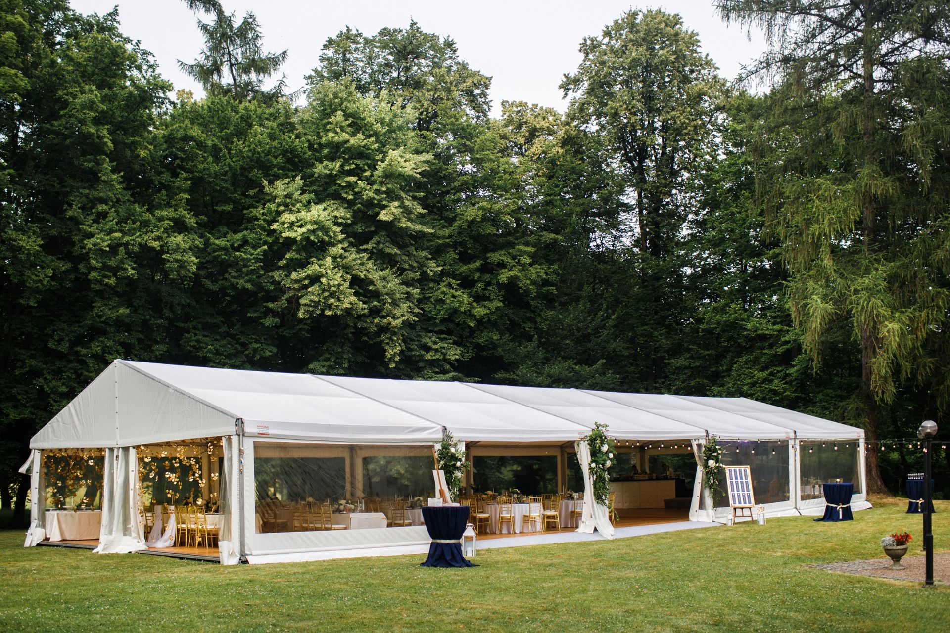 A large white event tent with transparent walls set up on a grassy lawn surrounded by dense green trees.