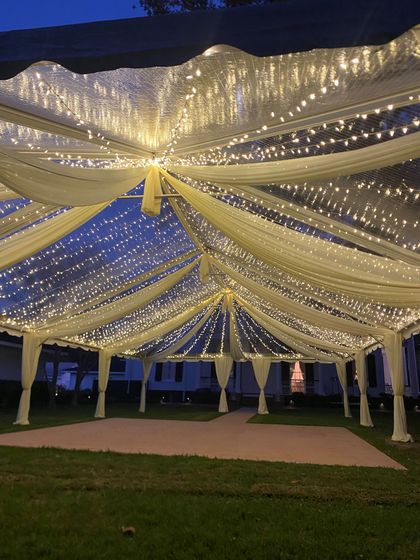 Draped white fabric canopy lit with warm string lights over a grassy outdoor event space at night Draped white fabric canopy lit with warm string lights over a grassy outdoor event space at night