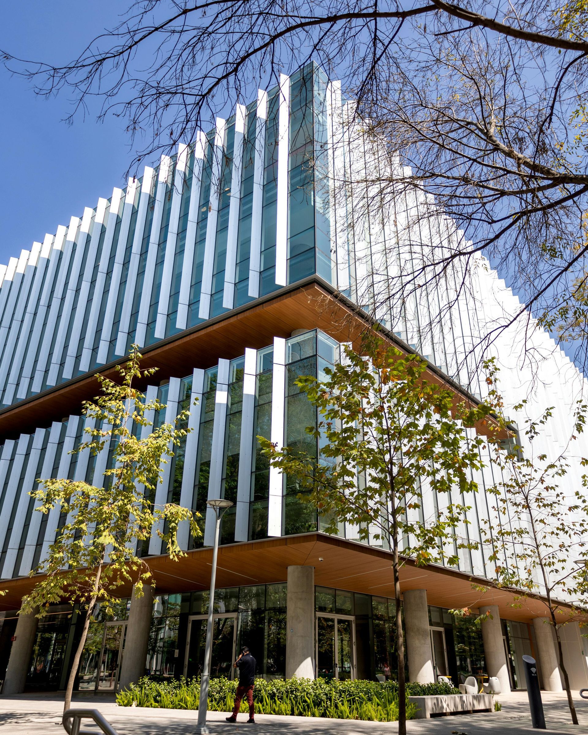 Modern building with white vertical accents, glass windows, and a brown overhang, partially obscured by trees.