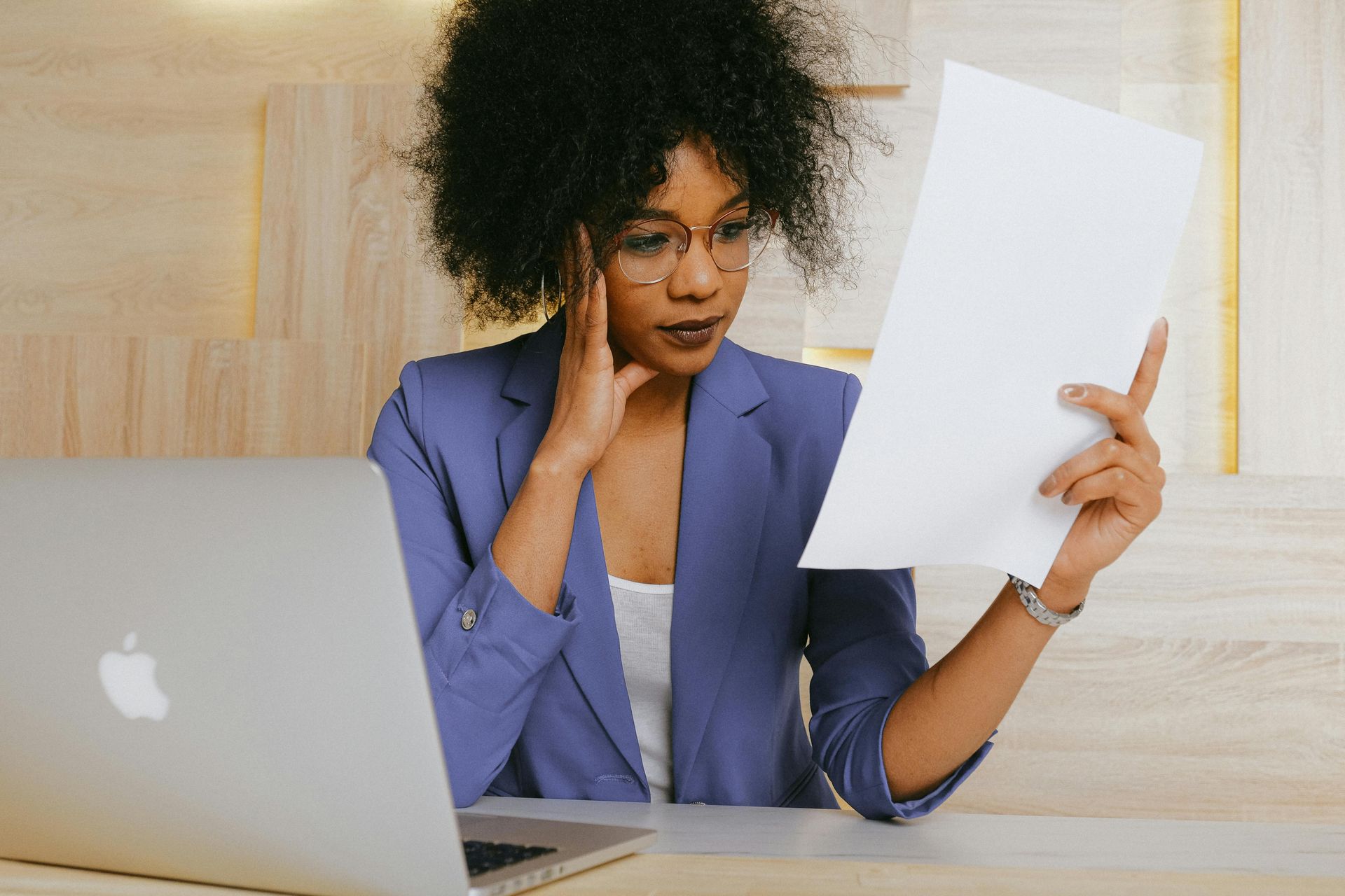 Woman in blue blazer, glasses, studying documents, laptop, appearing concerned.