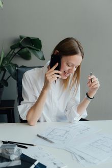 Woman in white shirt on the phone, smiling, looking at architectural drawings on a desk. Plant in background.