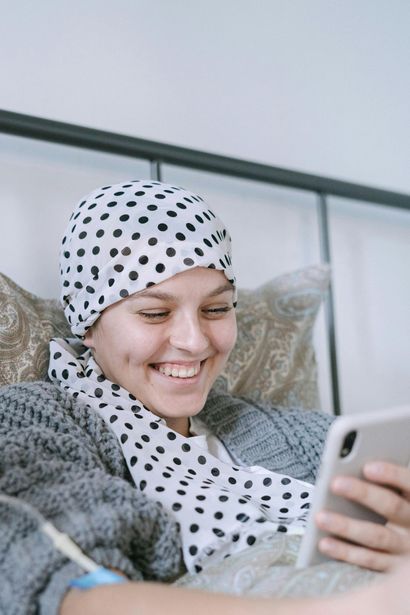 Person with headscarf smiles, looking at a phone while in bed. Intravenous drip in arm.