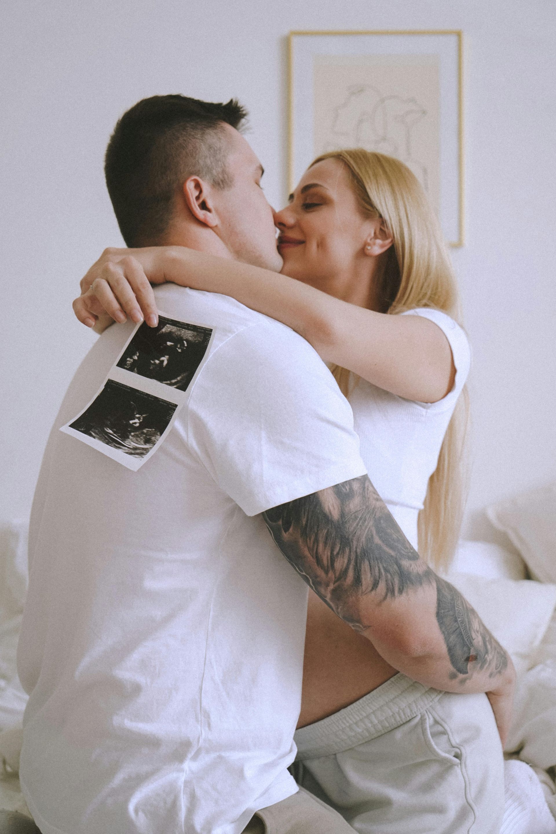 Couple embraces, kissing, holding sonogram photos. White room, soft light.