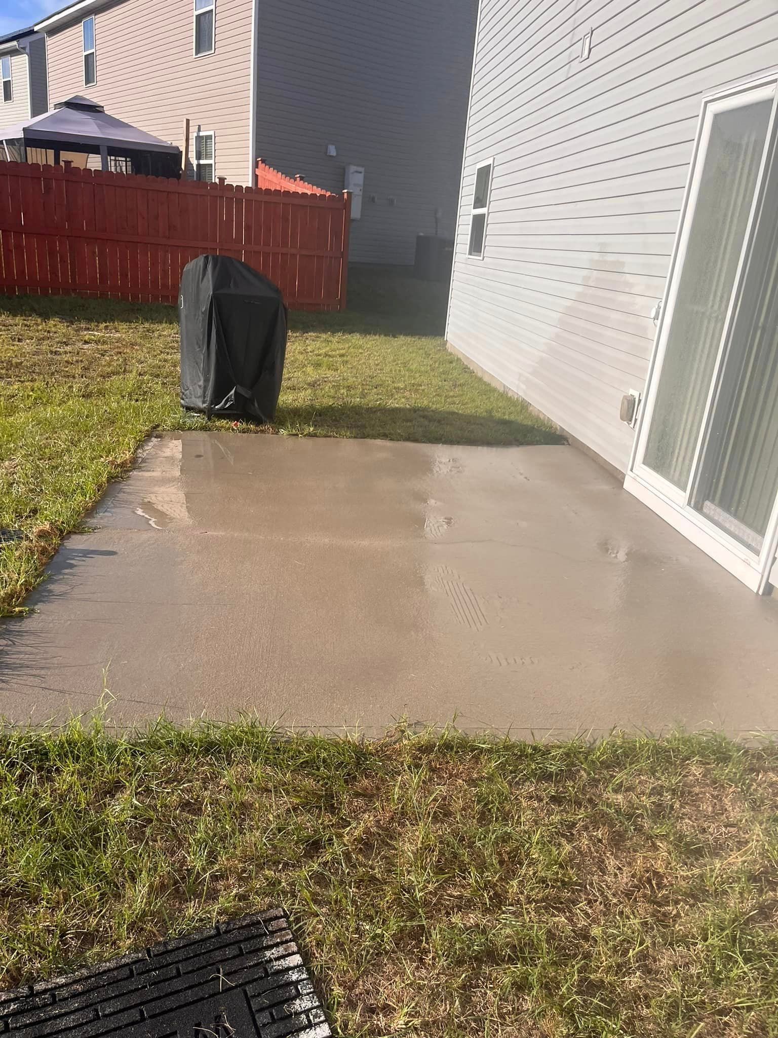 A patio in the backyard of a house with a grill and a fence.