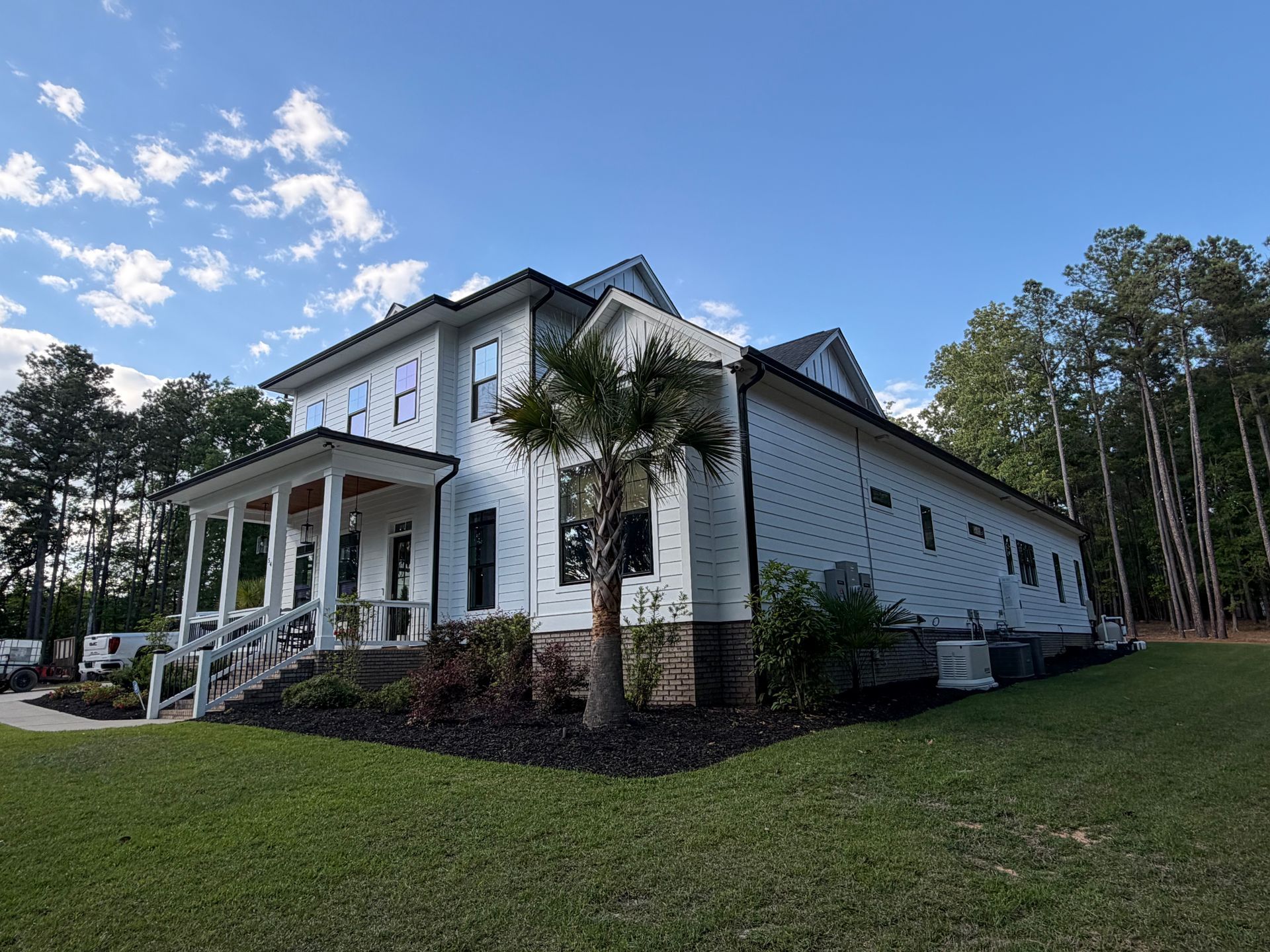 White two-story house with porch, black roof, and palm trees on a grassy lawn under a blue sky