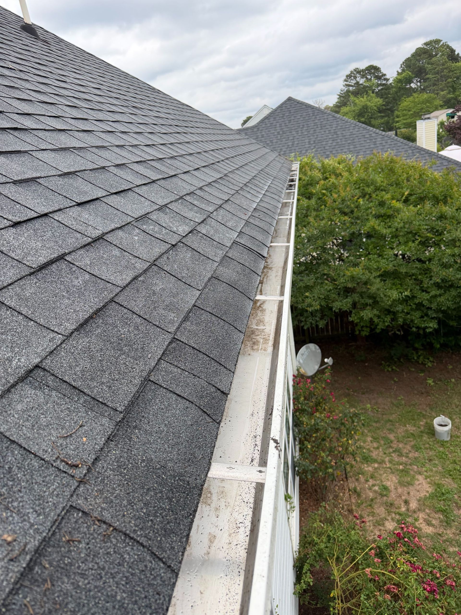Roof shingles and white gutter beside a green hedge and cloudy sky, viewed along a roofline