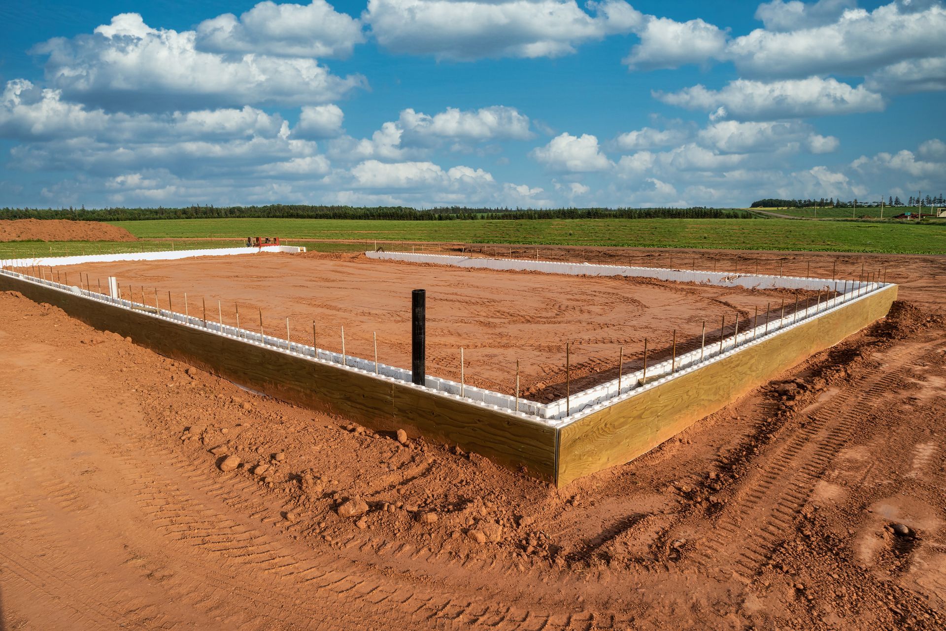 Construction site with a rectangular concrete foundation laid on red soil under a cloudy blue sky.