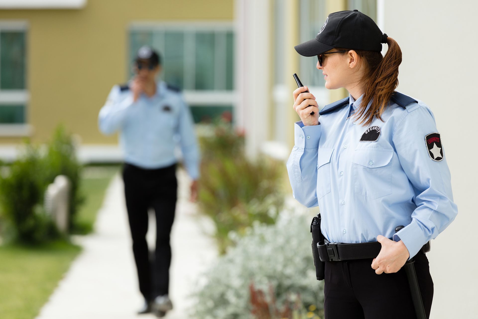 A woman is talking on a walkie talkie while a man walks in the background.
