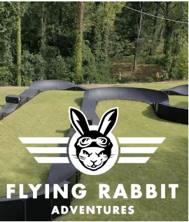 Flying Rabbit Adventures logo over a bike track. A bunny with goggles and wings on a white background. Green grass, black track, and trees in the background.