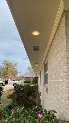 Exterior of a building with recessed lights and brick walls. Bushes and a street visible under cloudy sky.