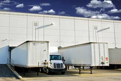 Trucks docked at a loading dock of a large white warehouse under a blue sky.