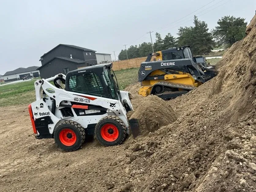 A Bobcat Is Driving Down a Dirt Road Next to A Bulldozer — Exeter, NE — Axline Landscaping & Trees