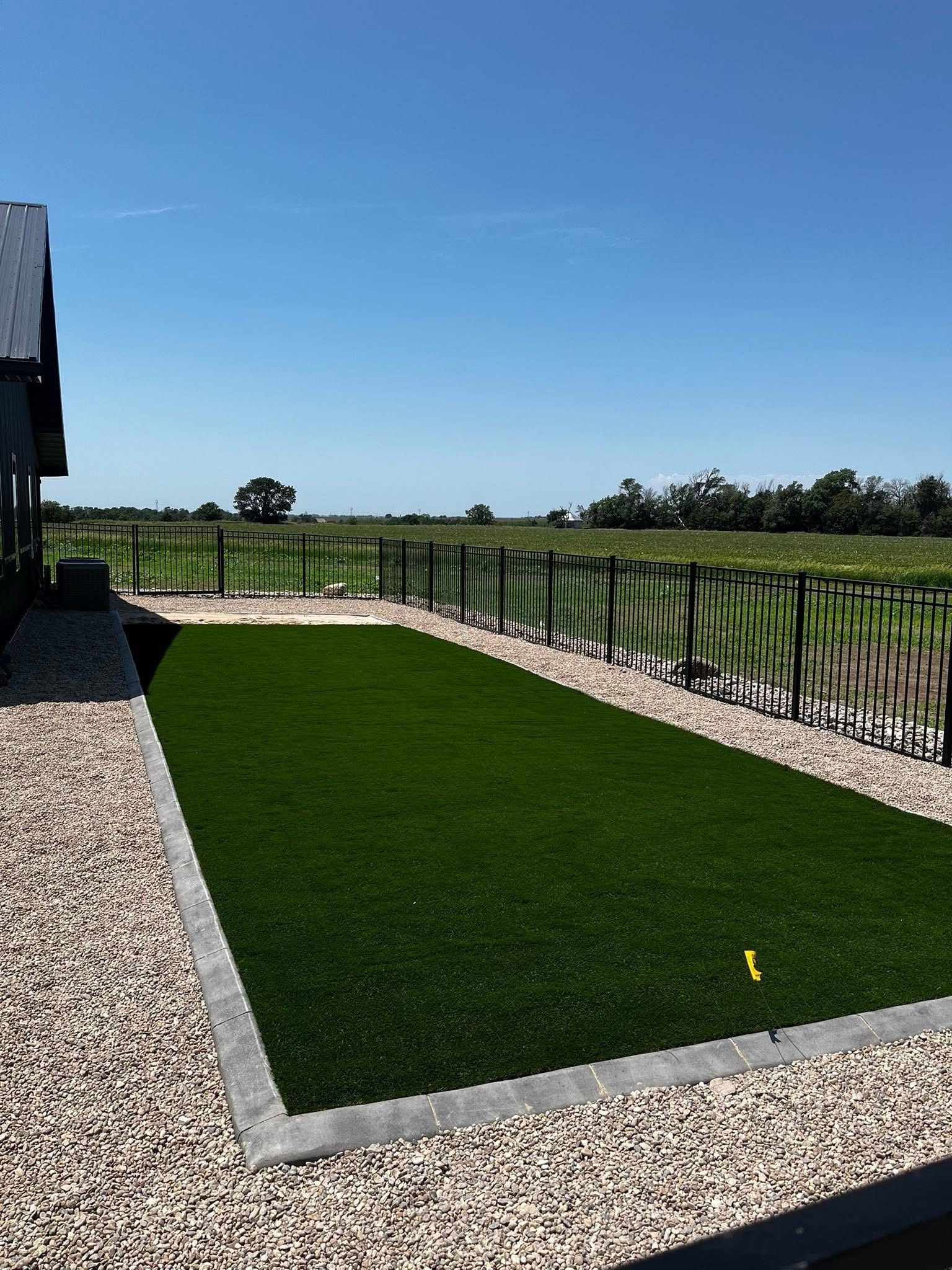 Green artificial turf surrounded by beige gravel and a black fence under a blue sky.