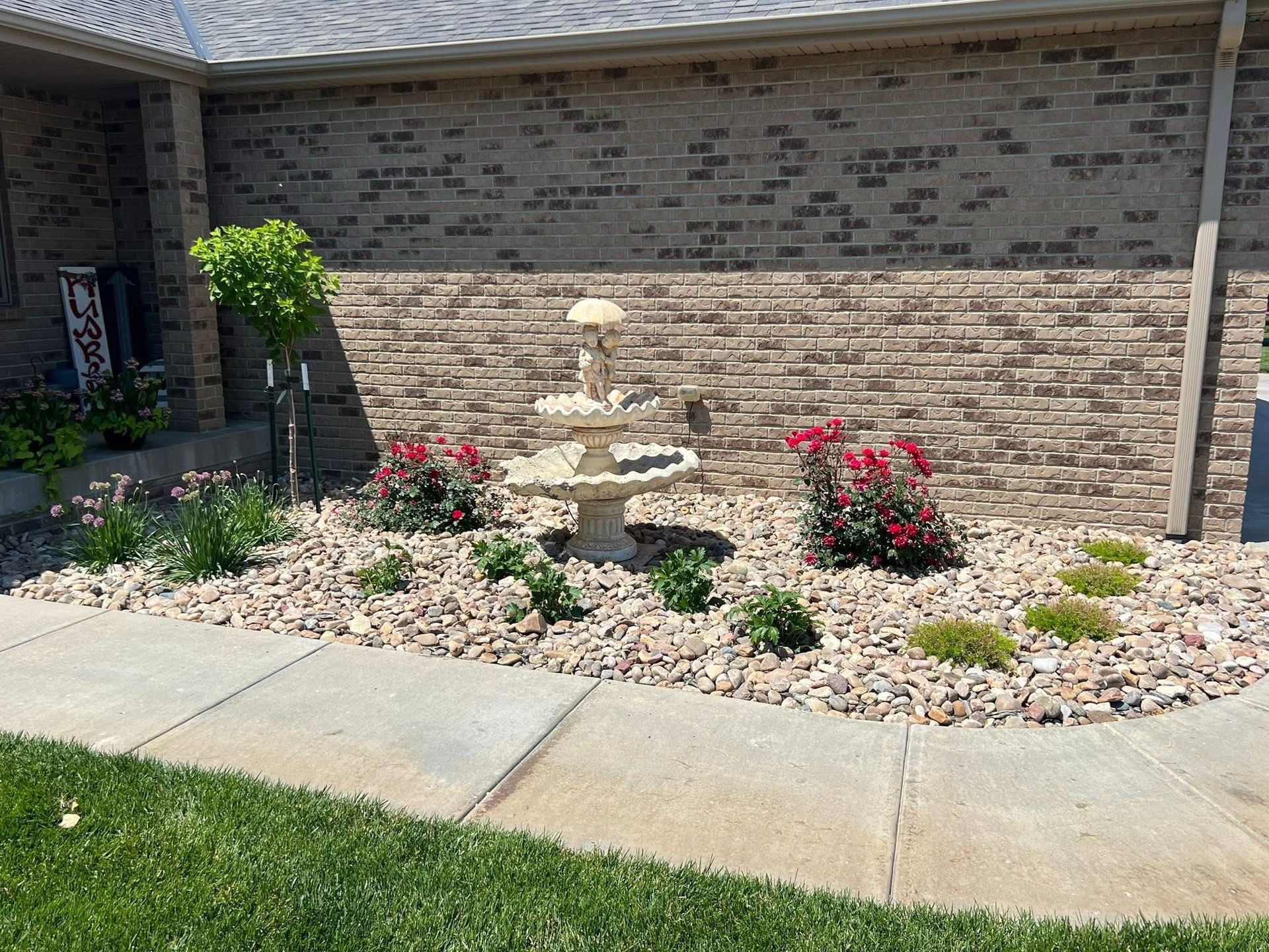 Front yard garden bed with stone, a tiered fountain, and red flowering plants against a brick wall.