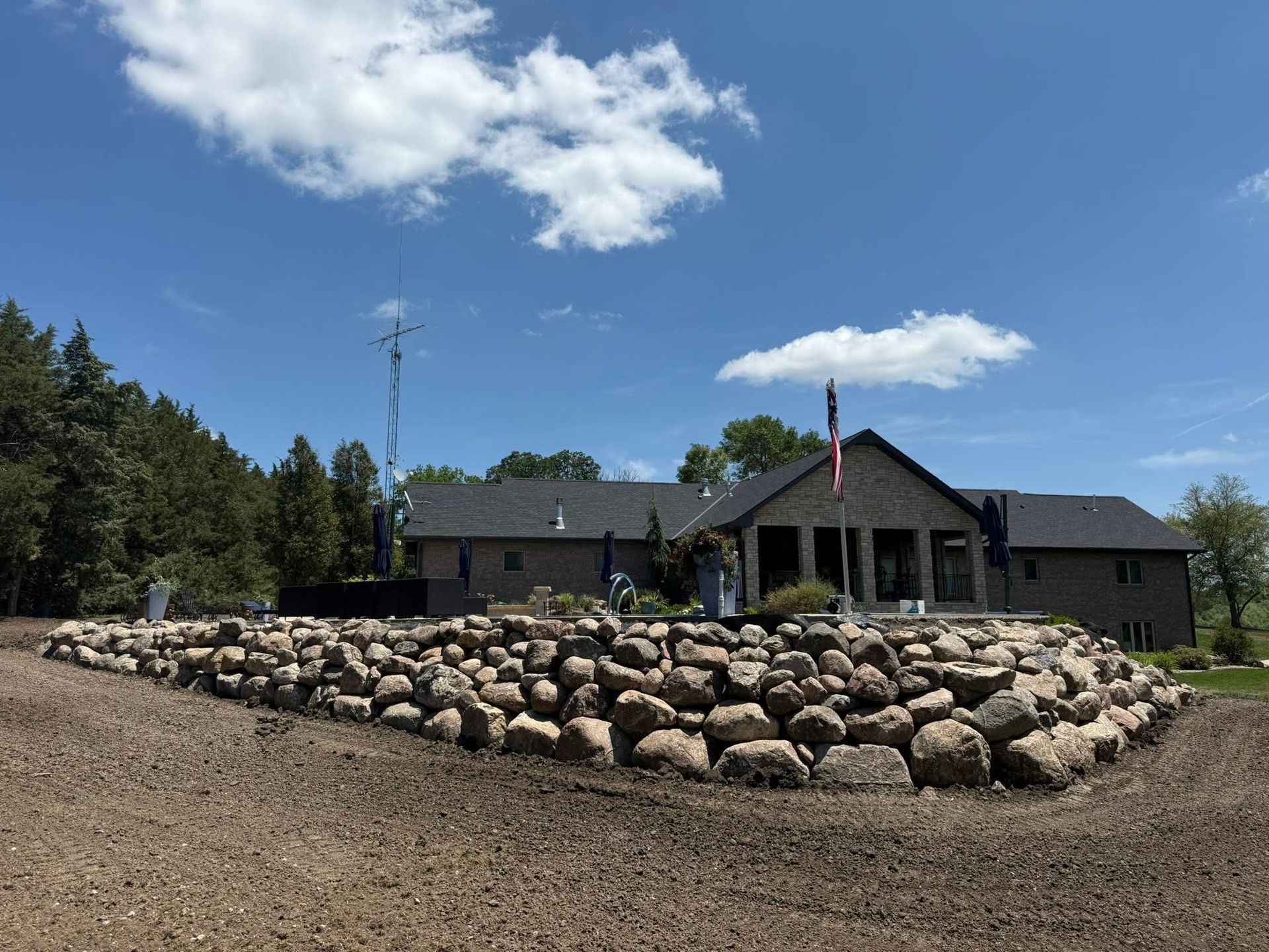 Brown stone house with rocky retaining wall, flag, and cloudy blue sky.