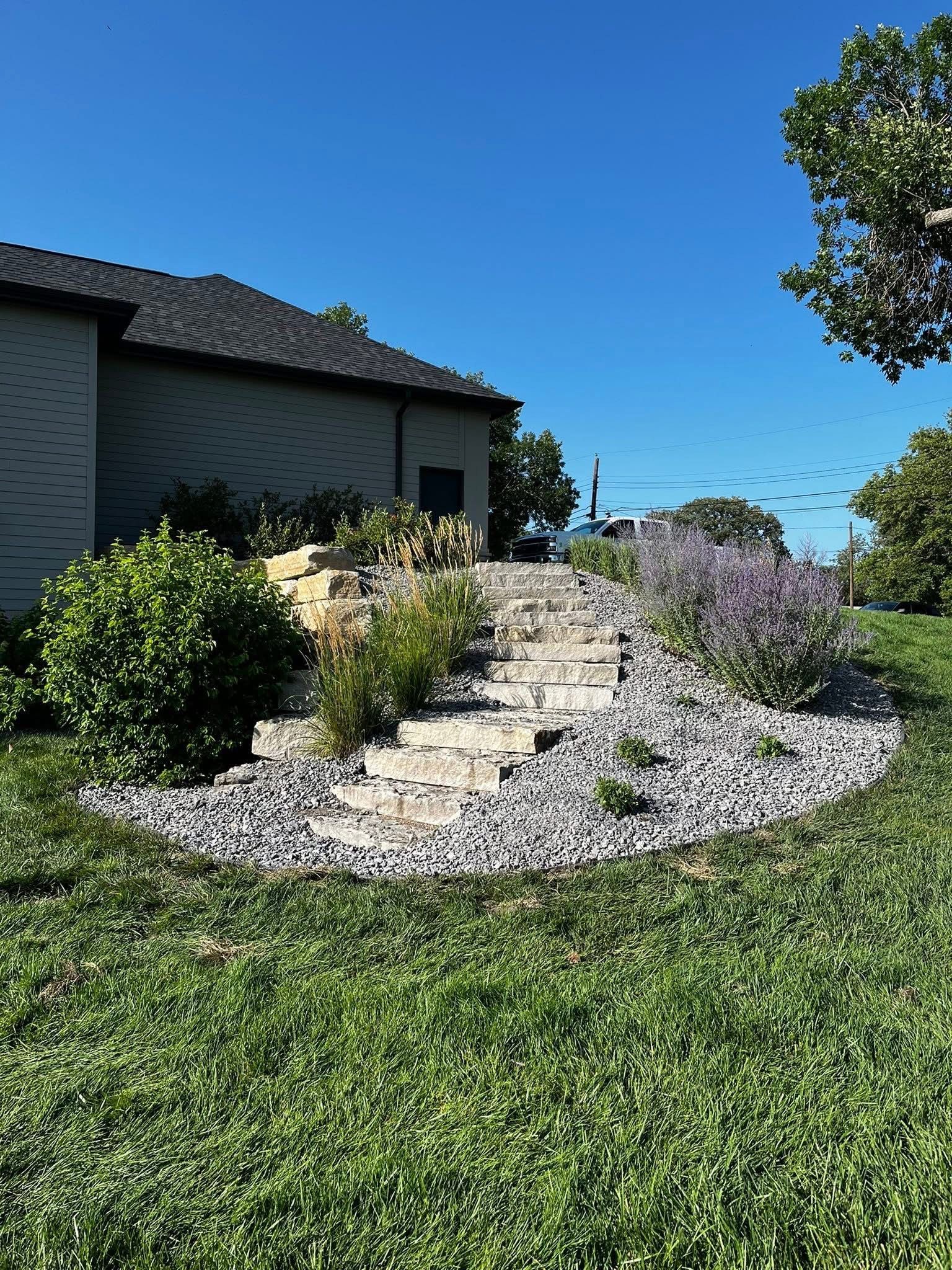 Stone steps lead up a landscaped area with plants and gray rocks; building in background, blue sky.