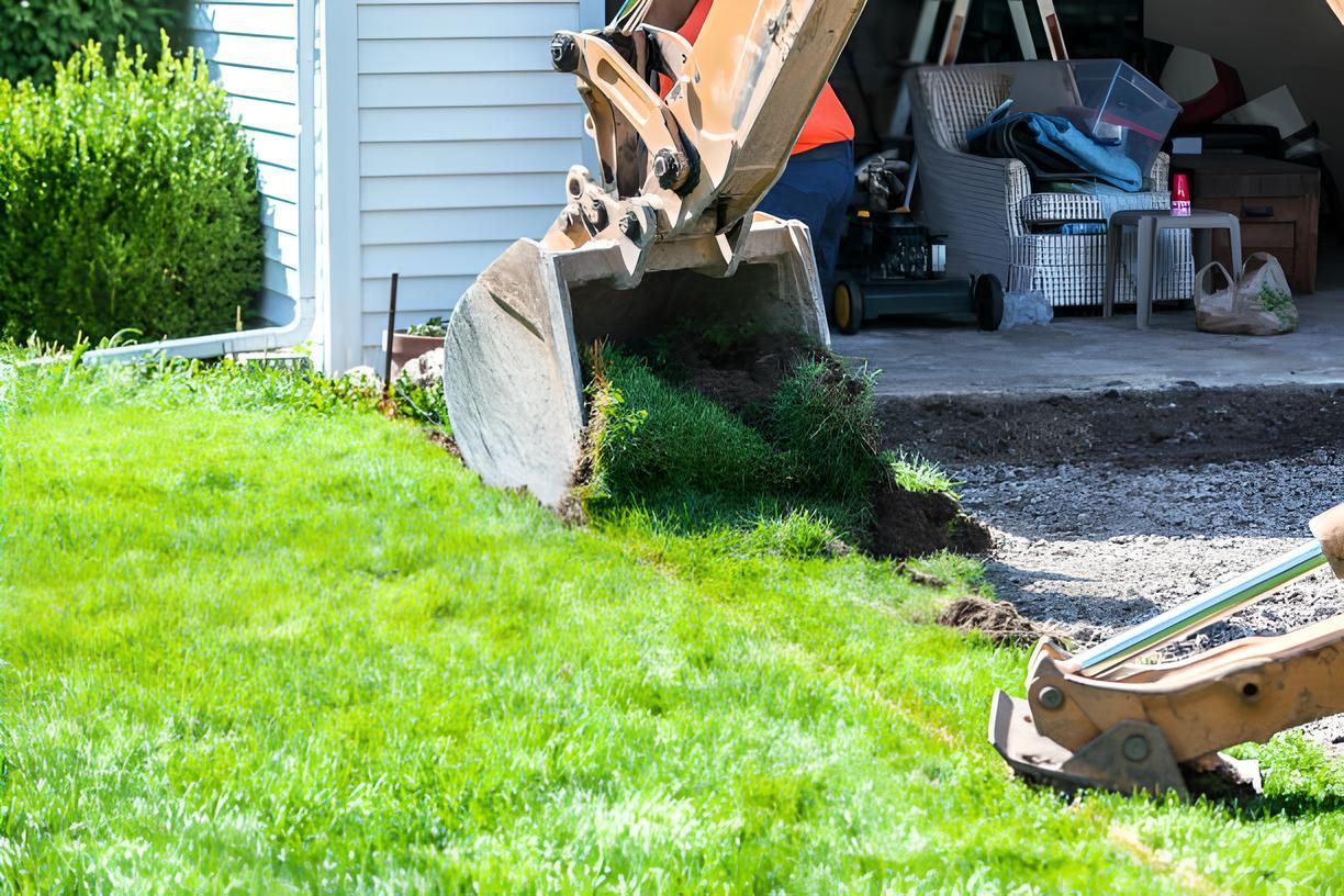 An excavator is digging a hole in the grass in front of a garage