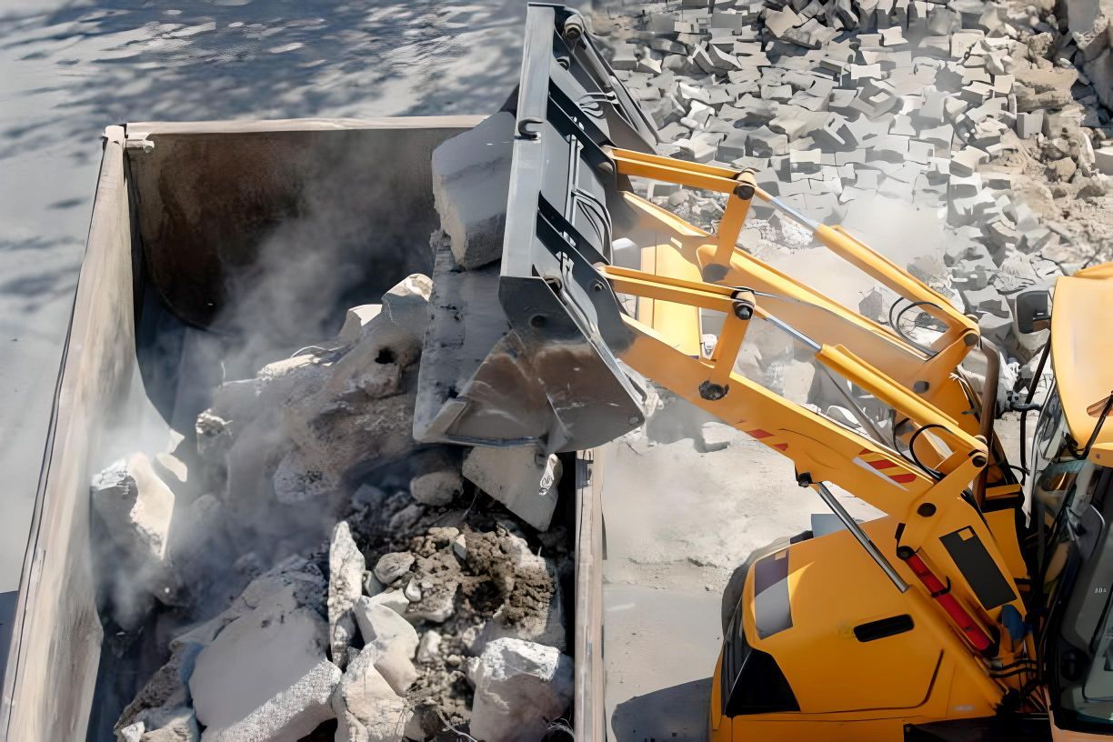 A yellow excavator is loading rocks into a dumpster