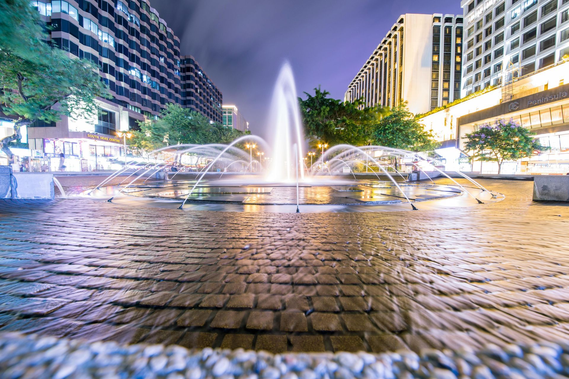 Fountain at night with water jets, cobblestone basin, and lit buildings in the background.