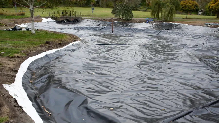 Black pond liner being installed in an excavated pond; white border on the edge, trees in the background.