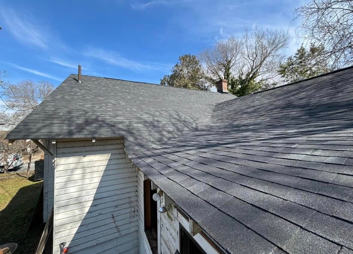 Dark gray asphalt shingle roof with light siding. Clear, blue sky with a few trees visible.