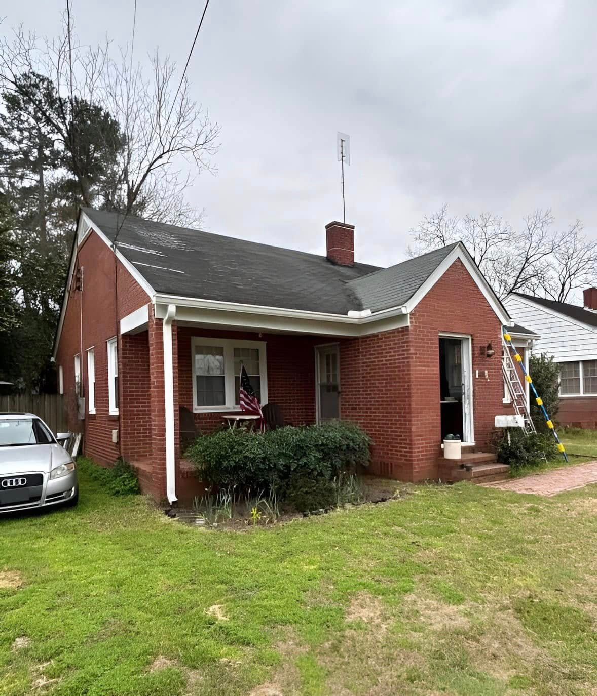 Red brick house with porch, green lawn, and a parked silver car. Overcast sky.