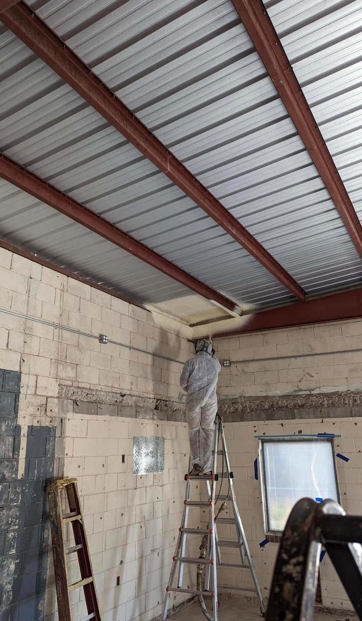 Person on ladder sprays insulation in a building with a metal ceiling and brick walls.