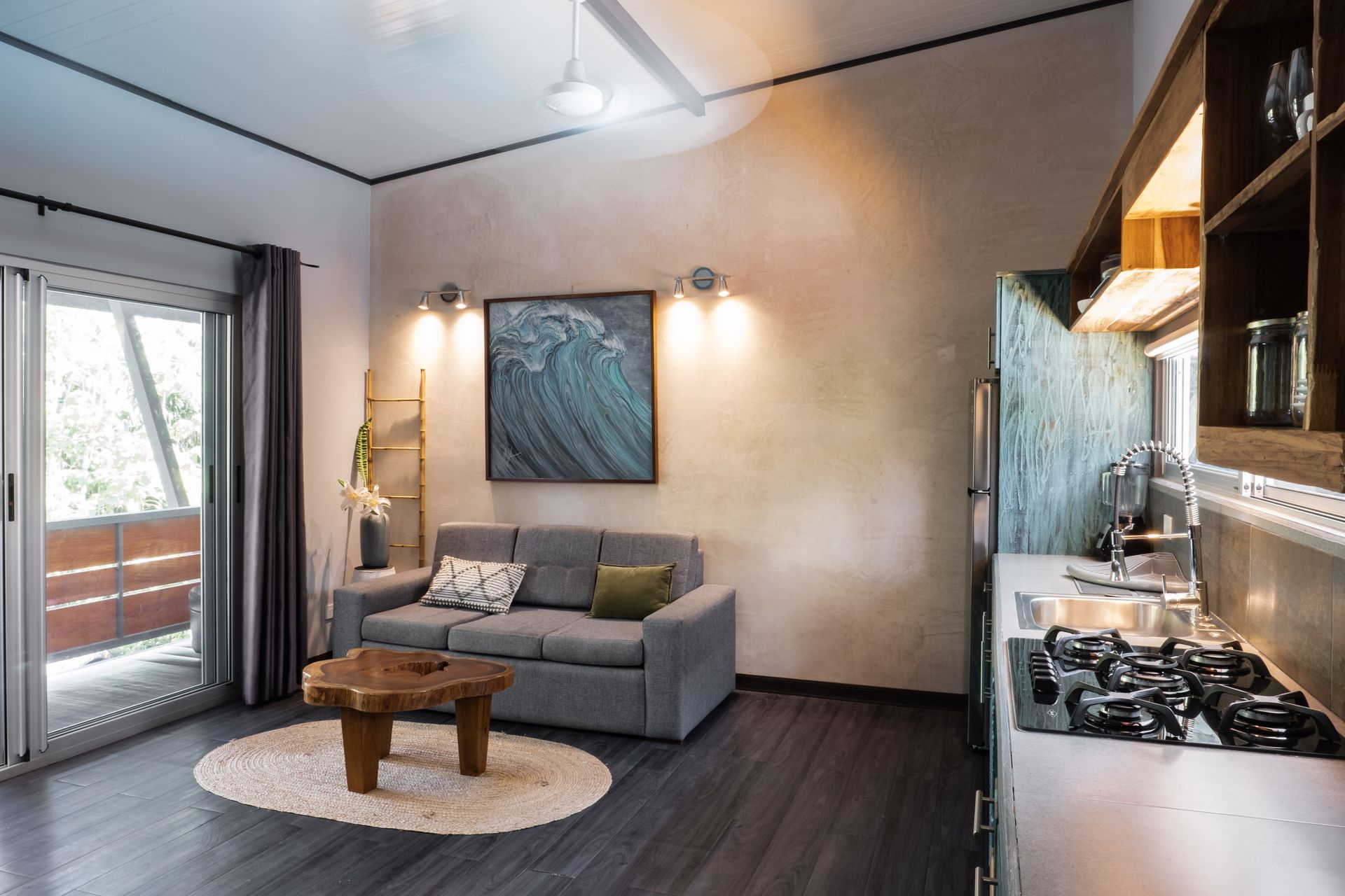A cozy studio apartment with a gray sofa, a wood coffee table on an oval rug, and a kitchen area with a gas stove.