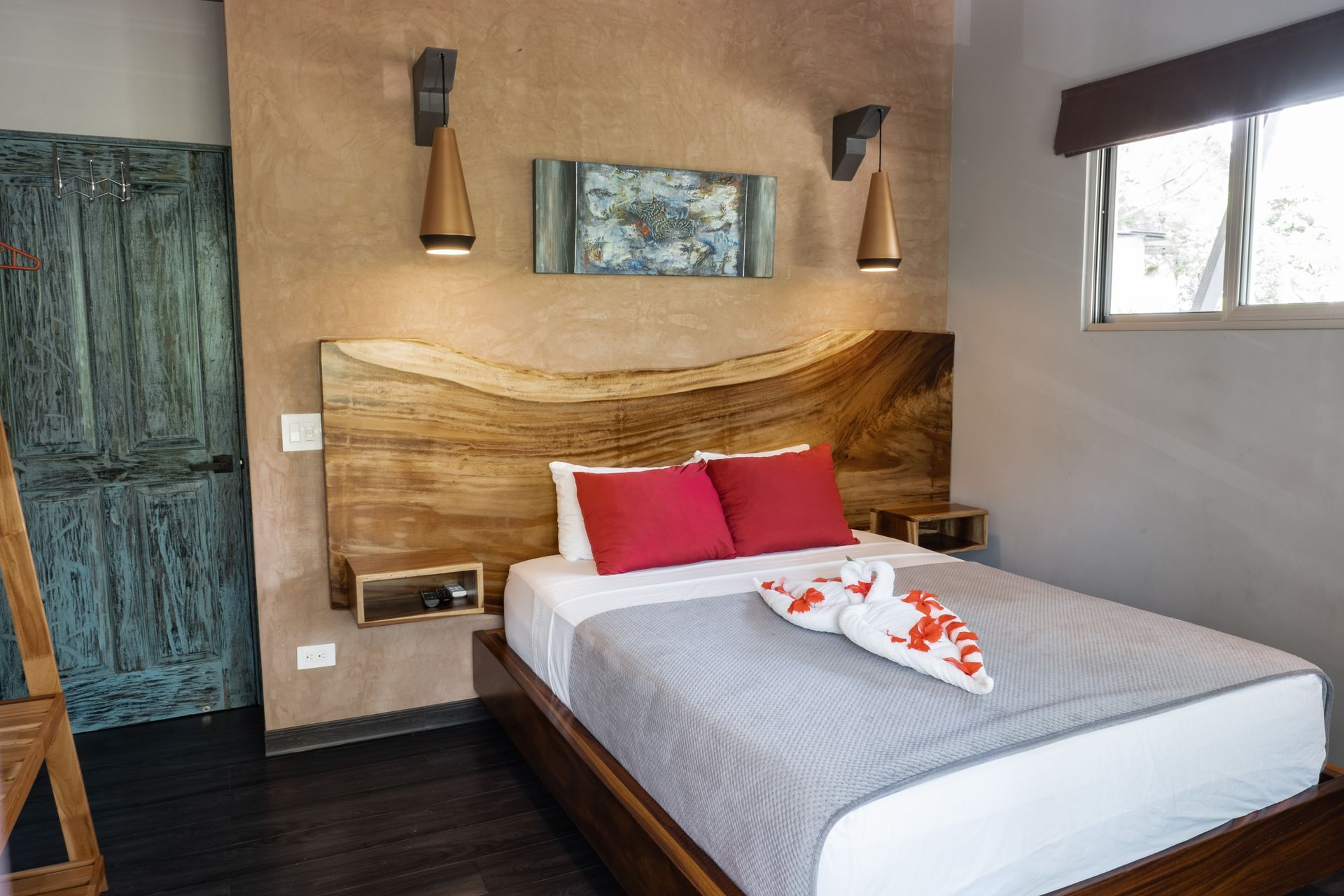 A hotel bedroom with a natural wood slab headboard, two red pillows, a grey bedspread, and two wall-mounted light fixtures.