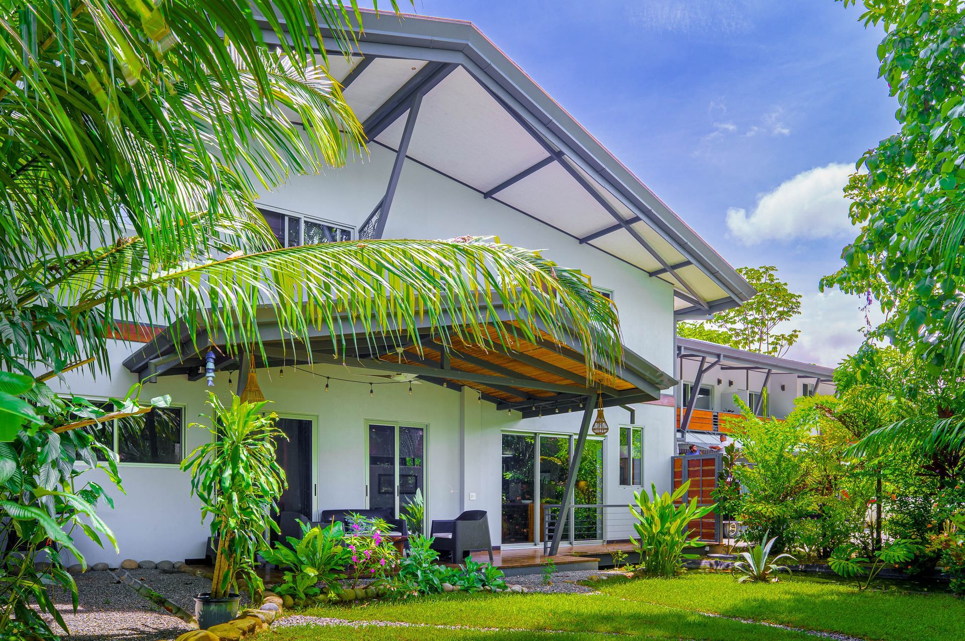 A two-story white house with a covered patio, surrounded by lush green foliage and trees under a clear blue sky.