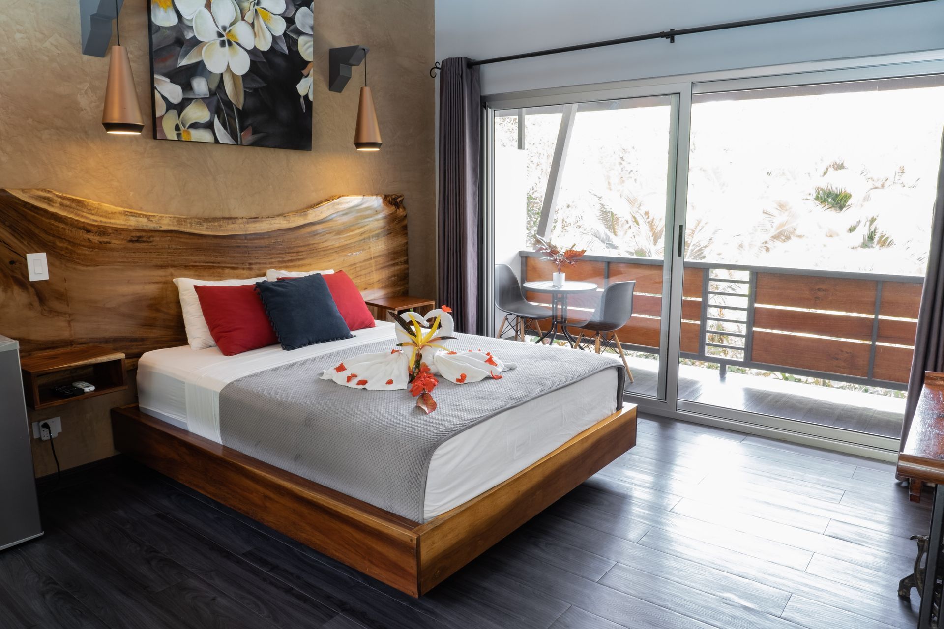 A modern hotel bedroom with a wooden headboard, a bed dressed with red and blue pillows, and a private balcony.