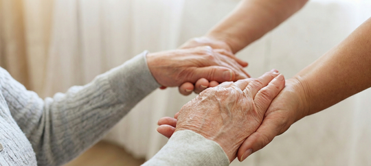 Person playing chess with an elderly man, smiling. Chessboard on a table, sunny room.
