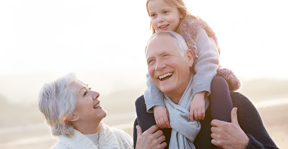 Grandfather carrying a child on his shoulders, grandmother smiling; outdoors, joyful.