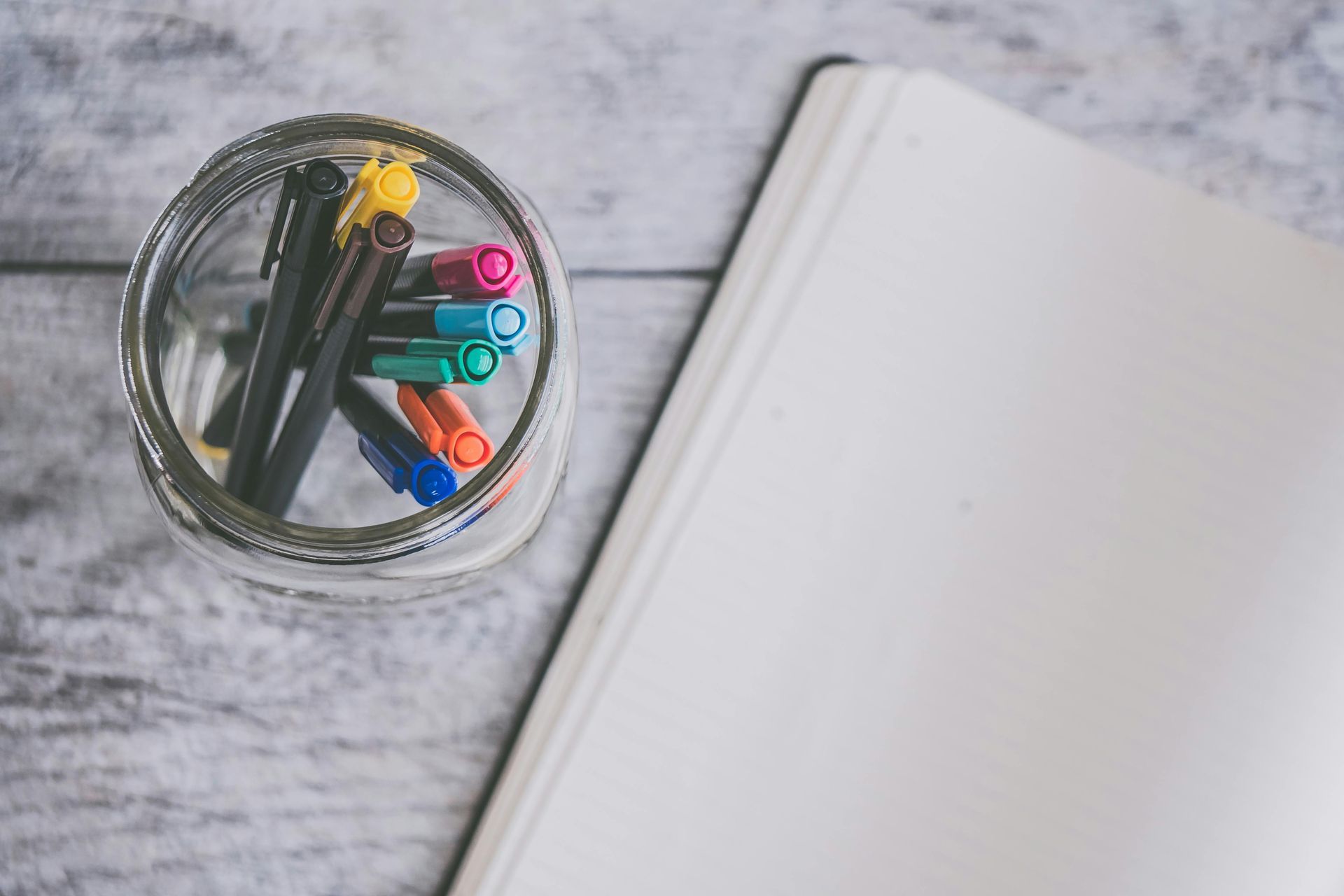 Jar of colorful pens next to an open notebook on a gray wooden surface.