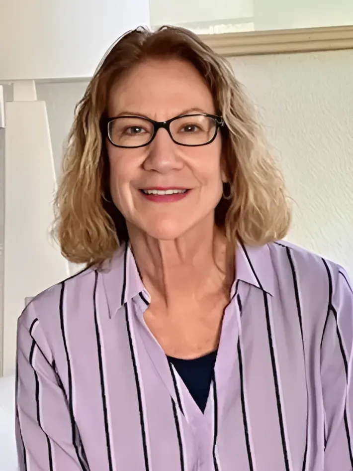 Woman with glasses smiles, wearing a striped purple shirt, indoors.
