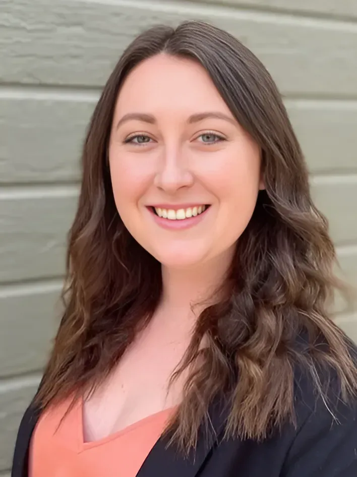Woman with brown hair smiles, wearing an orange top and black blazer in front of a wood-paneled wall.