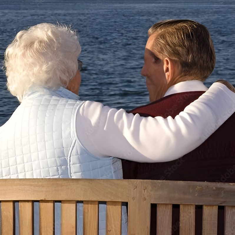 An elderly couple sits on a bench overlooking water, the man's arm around the woman.