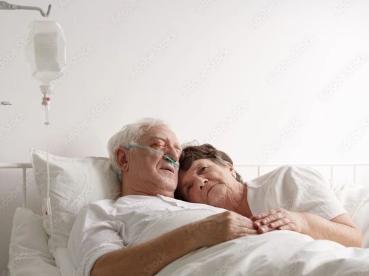 Elderly person in hospital bed with oxygen tubes, holding hands with a person, resting head on their shoulder.