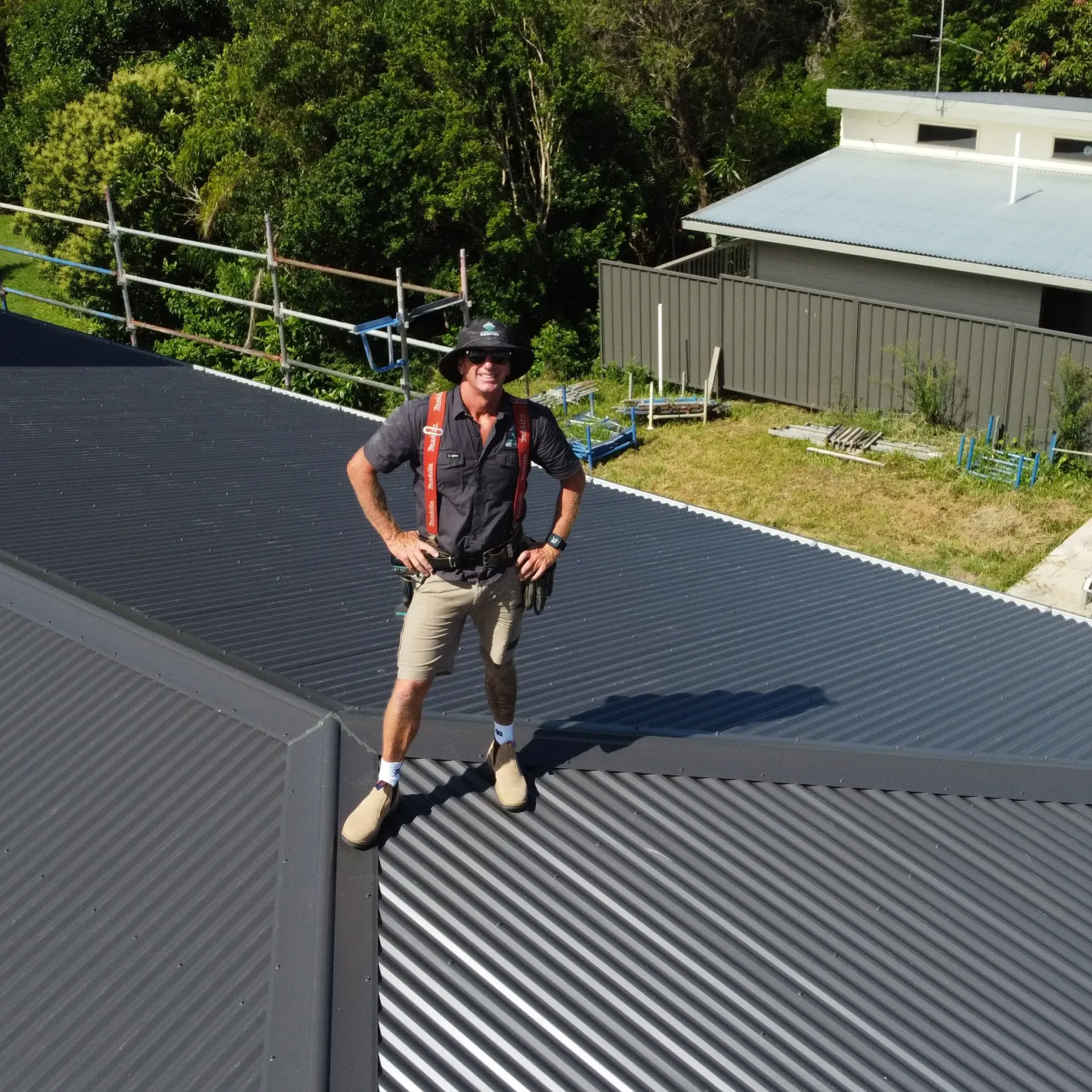 Man standing on a corrugated metal roof, arms at his sides, wearing a hat and work clothes.