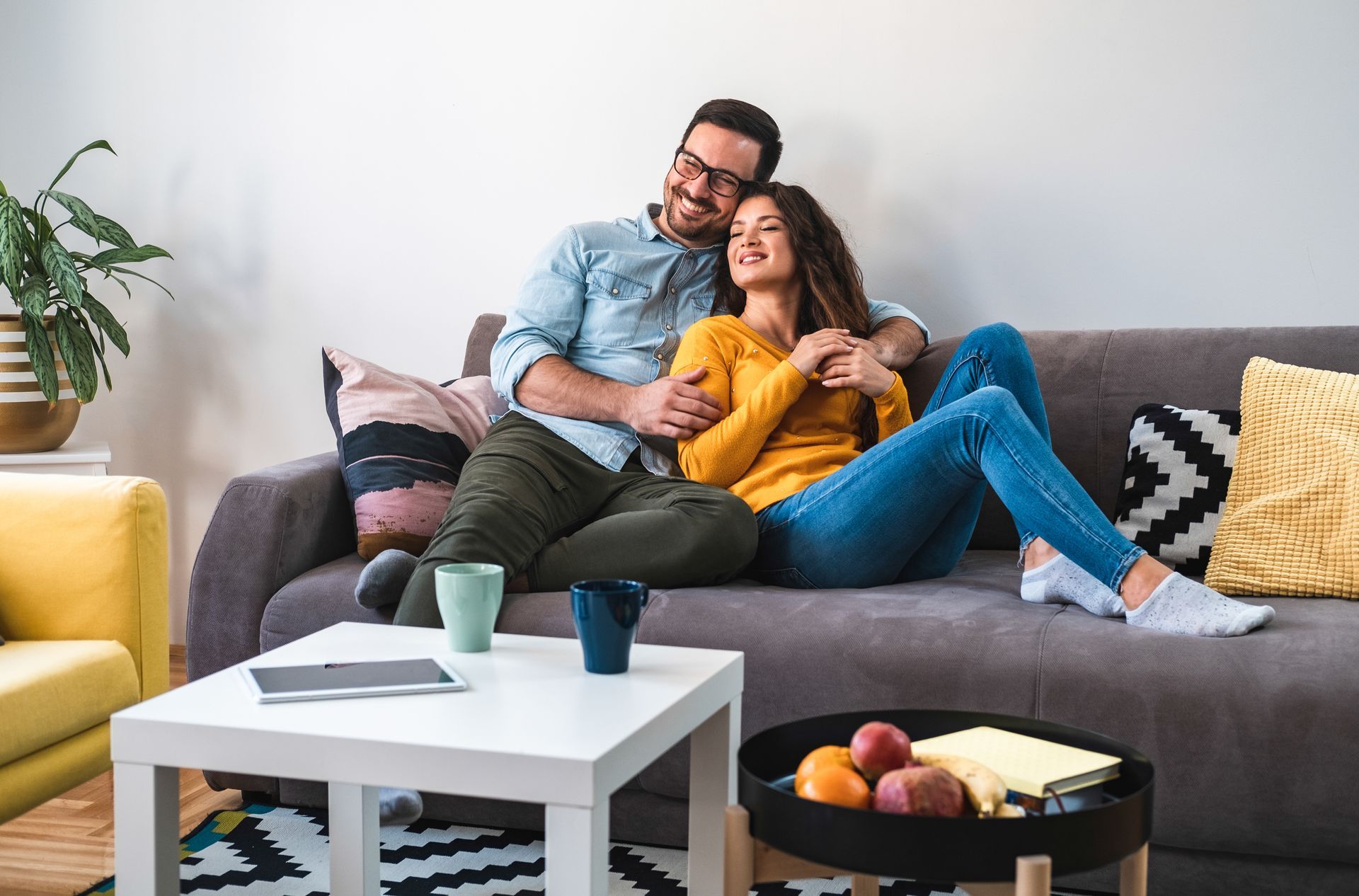 Couple cuddling on a gray couch in a bright living room with a coffee table and houseplants.