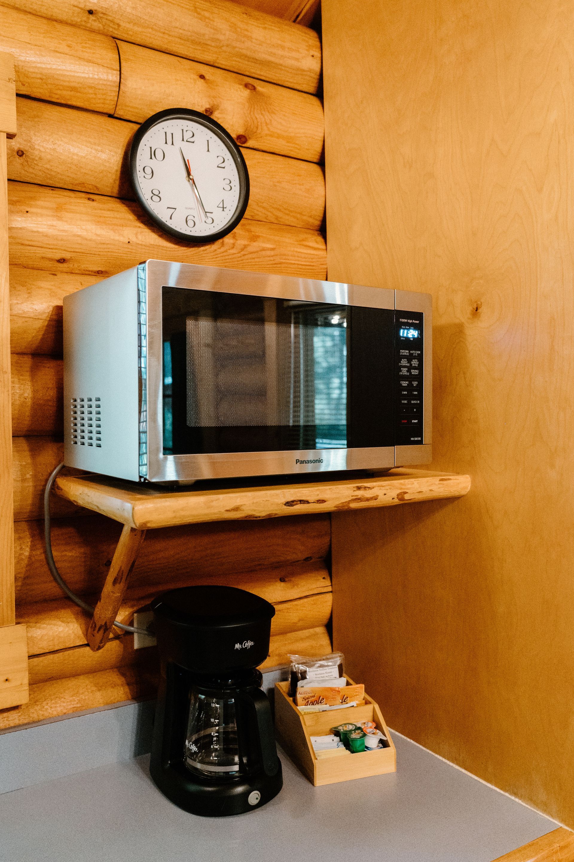 A microwave is sitting on a wooden shelf next to a coffee maker and a clock.