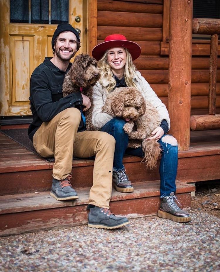 A man and a woman are sitting on the steps of a log cabin with their dogs.