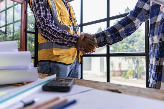 Two construction workers are shaking hands in front of a window.